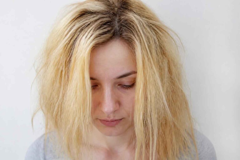 Kobieta ze spuszczoną głową i potarganymi blond włosami na jasnym tle